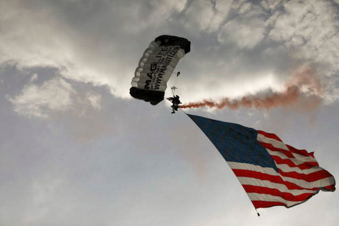 A soldier freefalls with a parachute while carrying an American flag and a flare leaves behind a trail of red smoke.