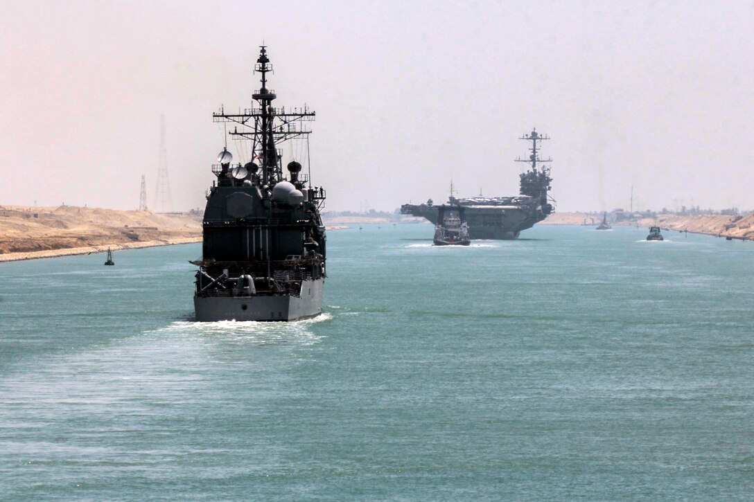 Multiple military ships move through a narrow passage of water.