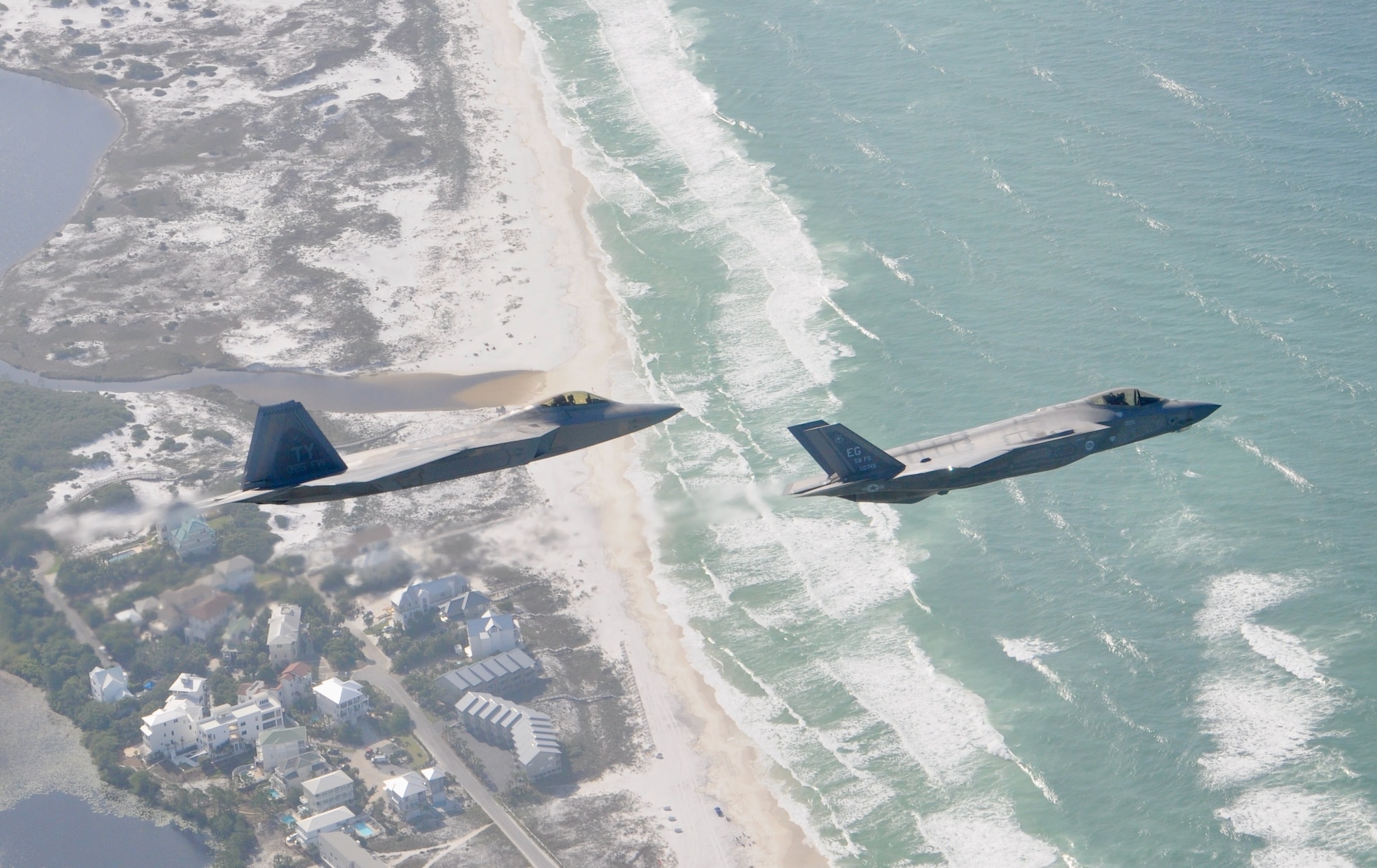 A F-22 Raptor and a F-35 Lightning II fly over the beach