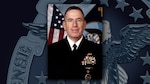 White man in dress uniform with medals and ribbons in front of the US flag.