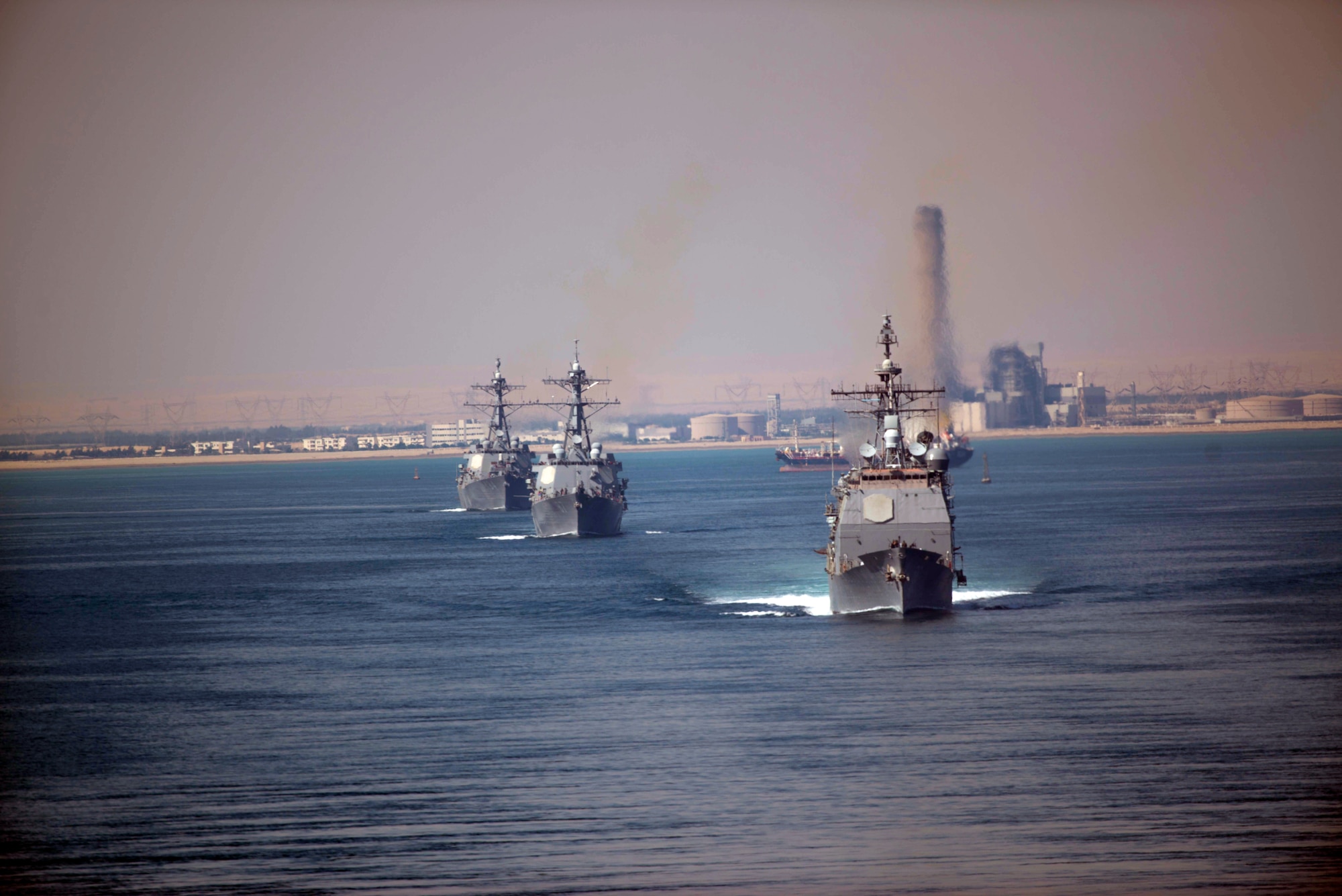 Three ships move through the water. In the rear is an industrial area.