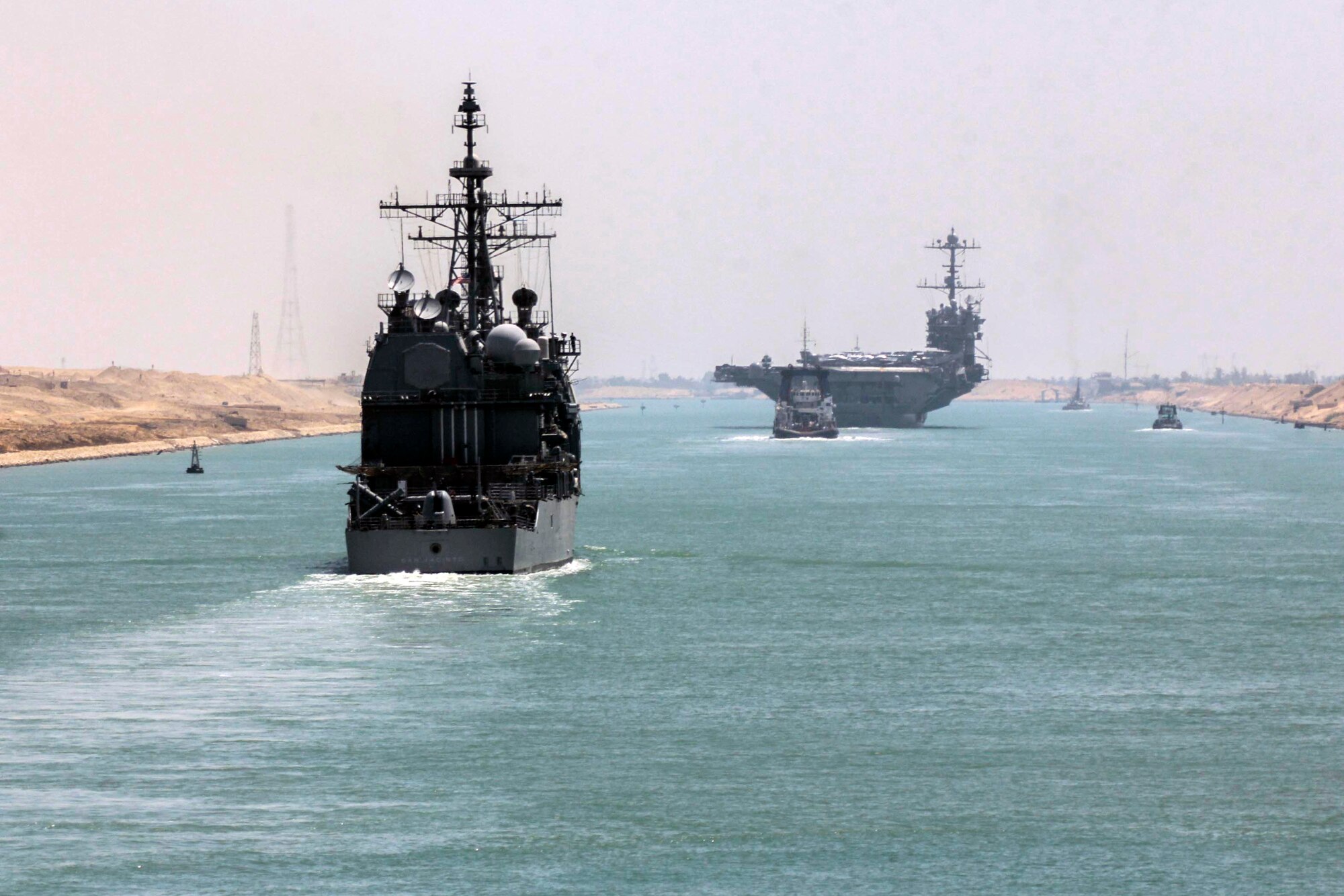 Multiple military ships move through a narrow passage of water.