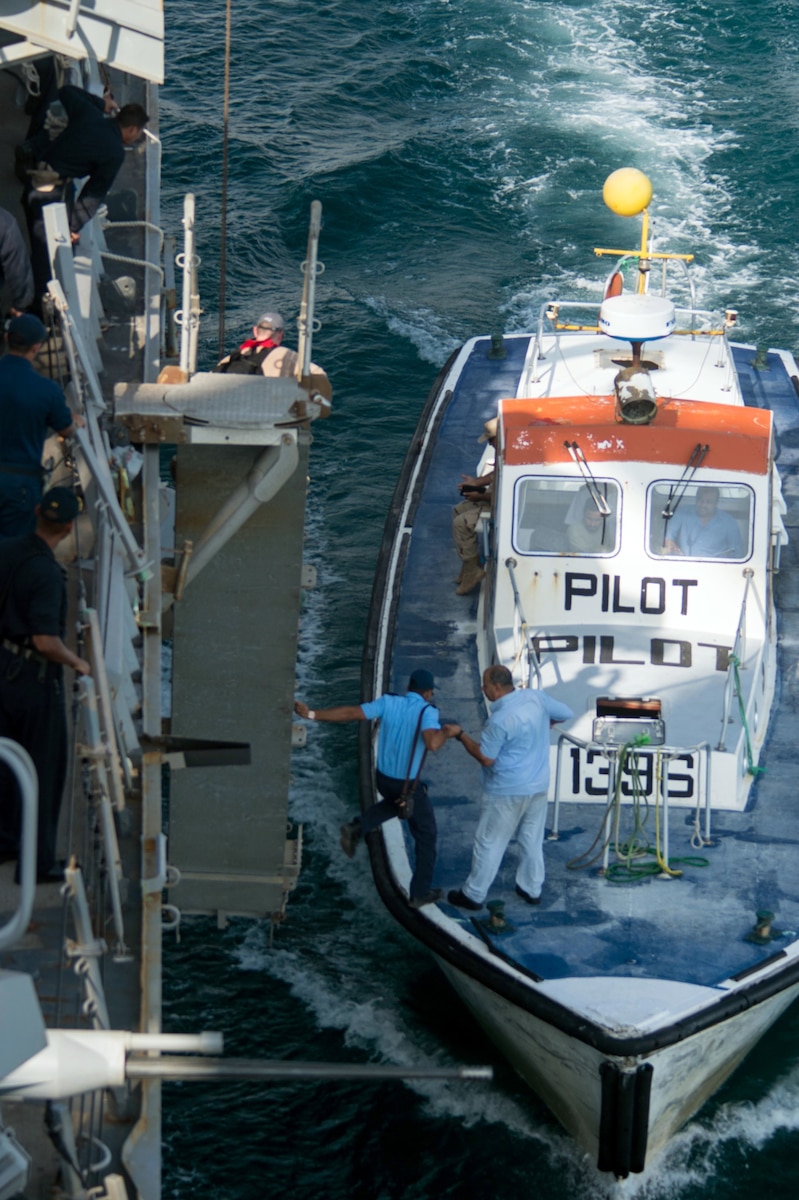 A small boat, with the word "pilot" written on the front, pulls alongside a larger vessel. Men move between the vessels.