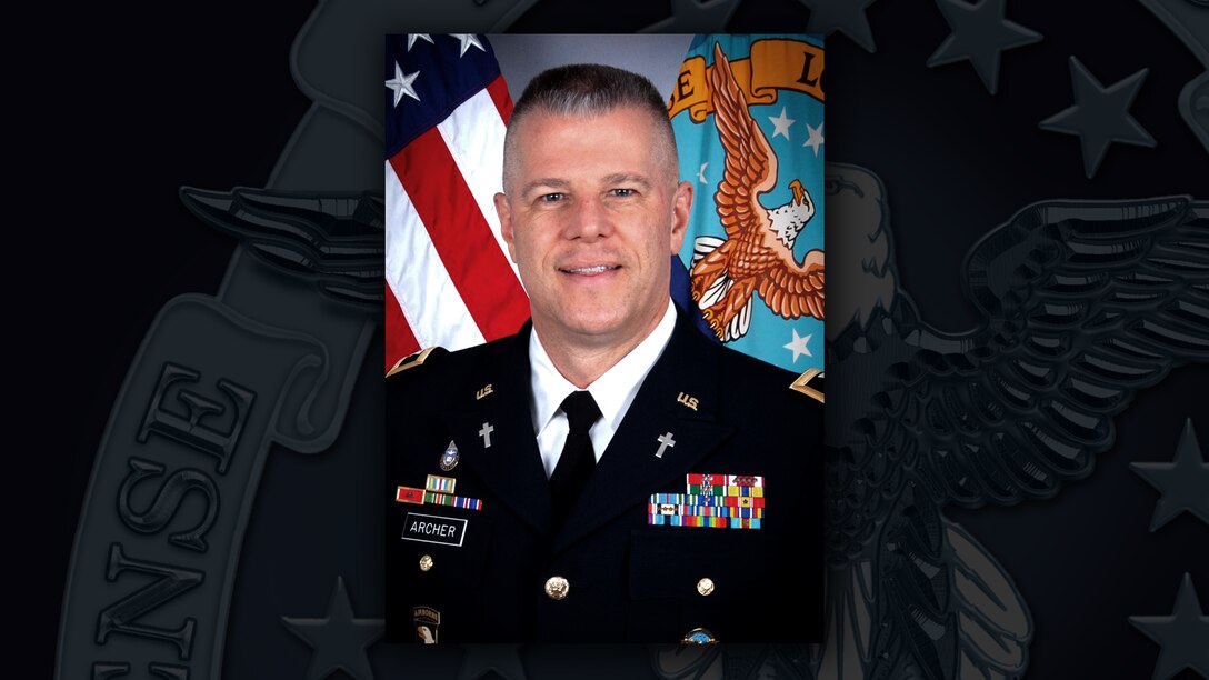 White man in dress uniform with medals and ribbons sits in front of US and DLA flags.