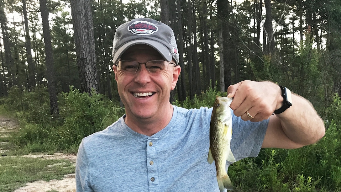 White man in baseball cap and grey shirt holds up a fish outdoors.