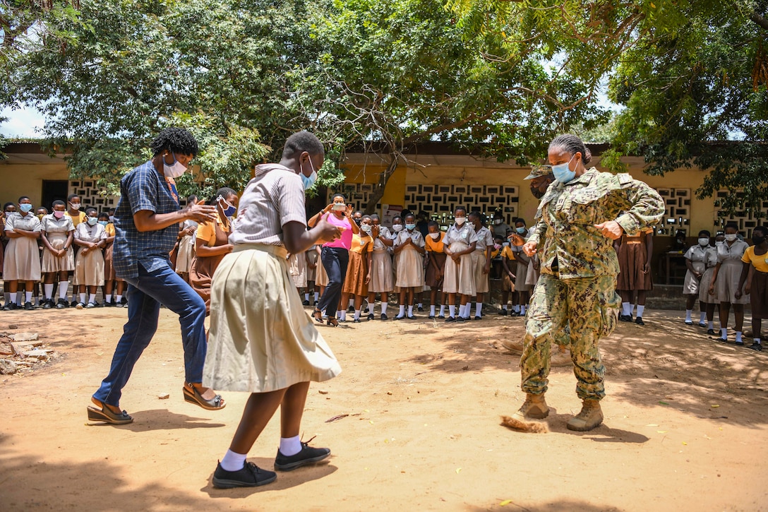 A service member dances with two Ghanaians on a dirt playground.