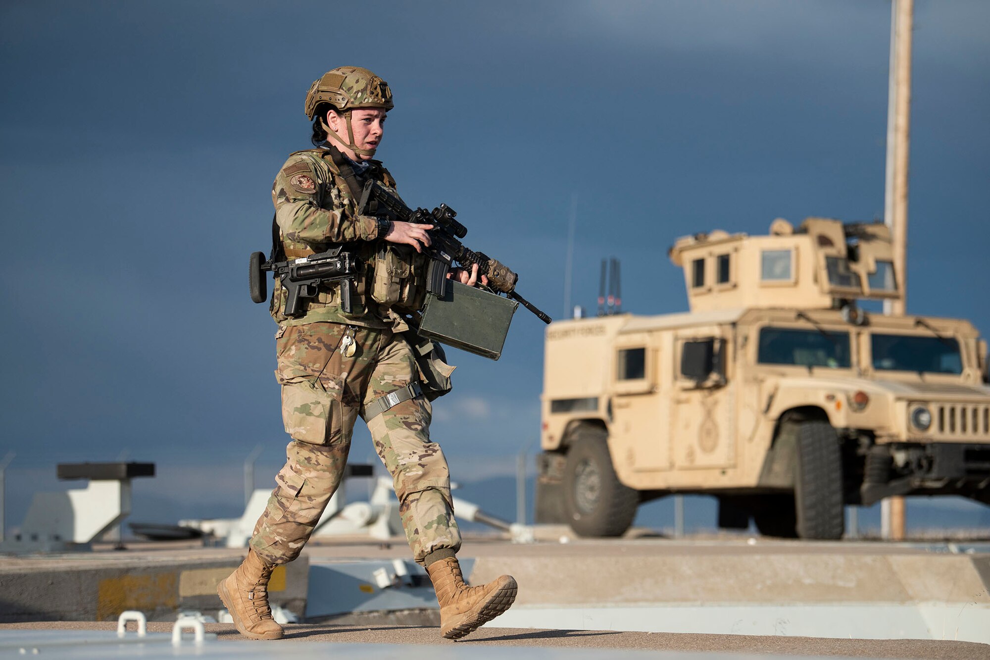 A female defender moves across the launch facility carrying a rifle and an ammo container, with a Humvee in the background.