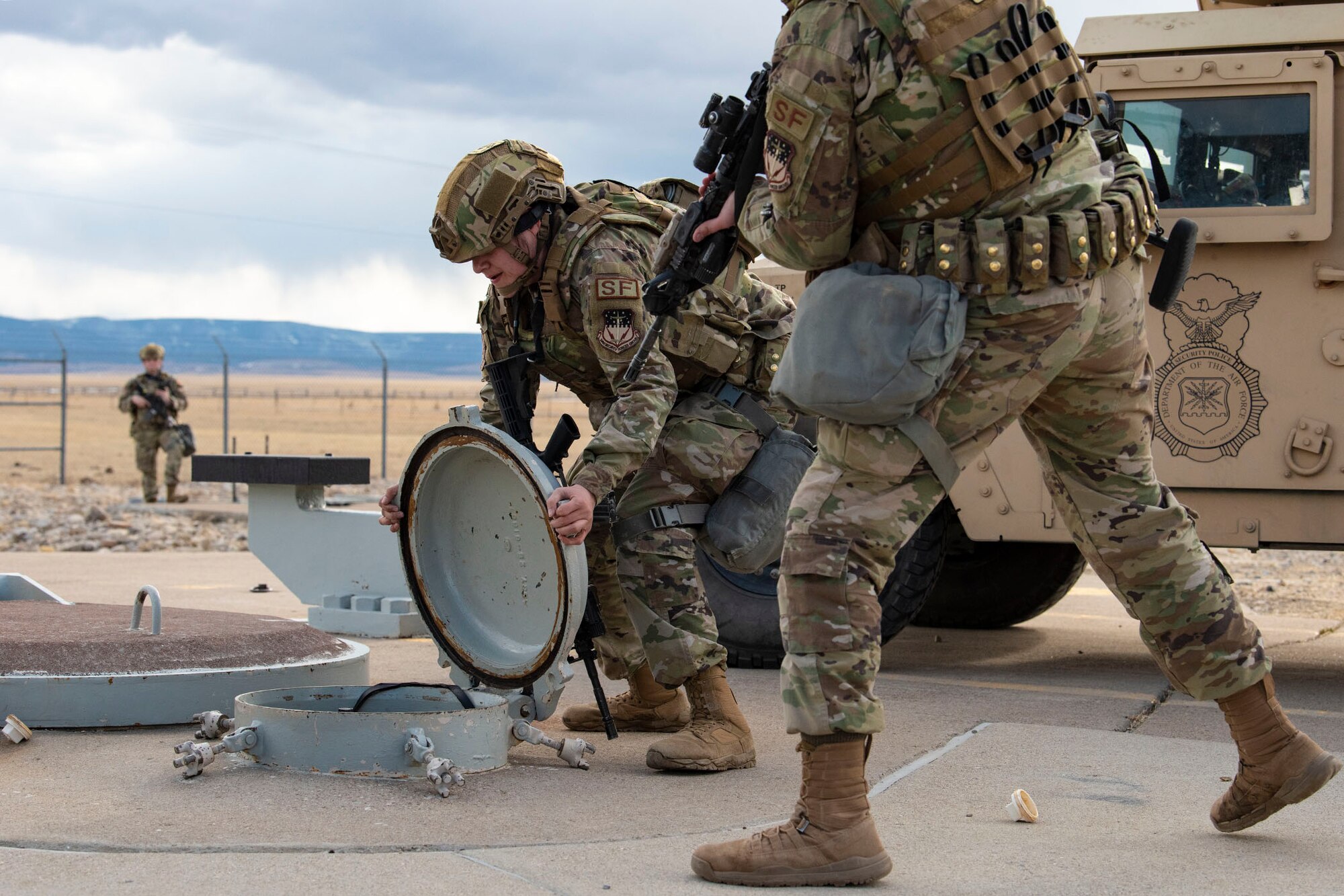 Defender opens a capsule and looks inside for explosive devices while another defender stands next to her with a rifle and a third defender is in the background of the frame holding a rifle and moving towards the open capsule.