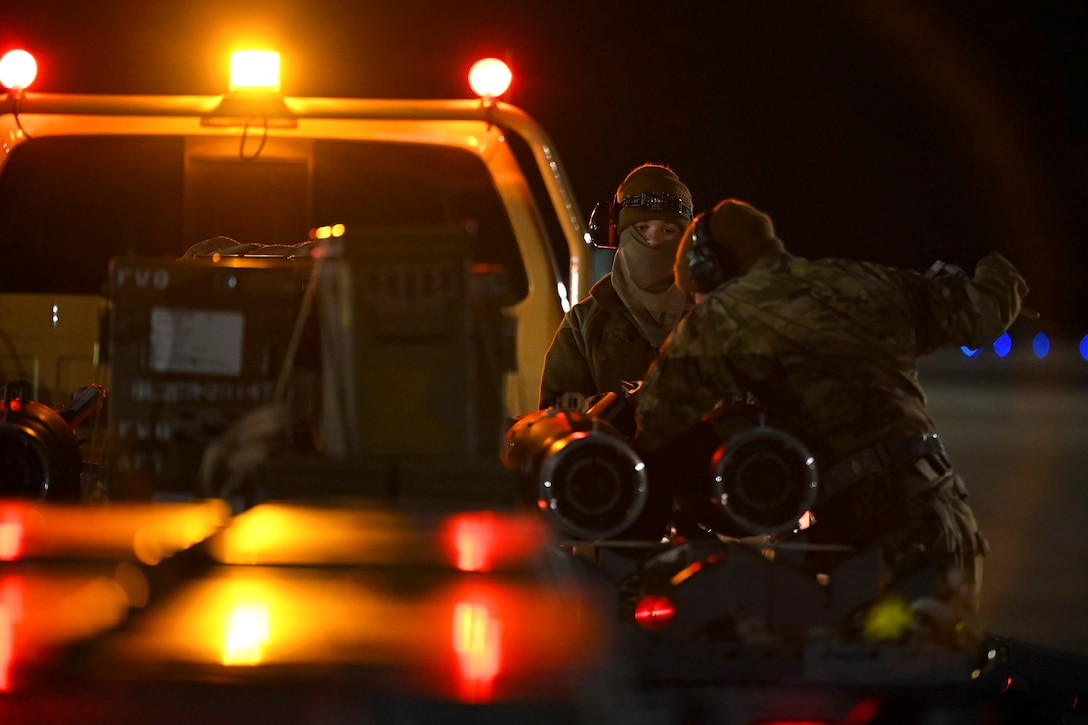 Airmen stand next to a vehicle in the dark illuminated by lights.