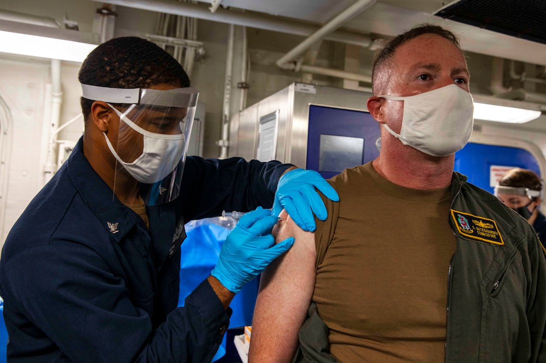 A sailor wearing a face mask and gloves gives a vaccine to a person wearing a face mask.