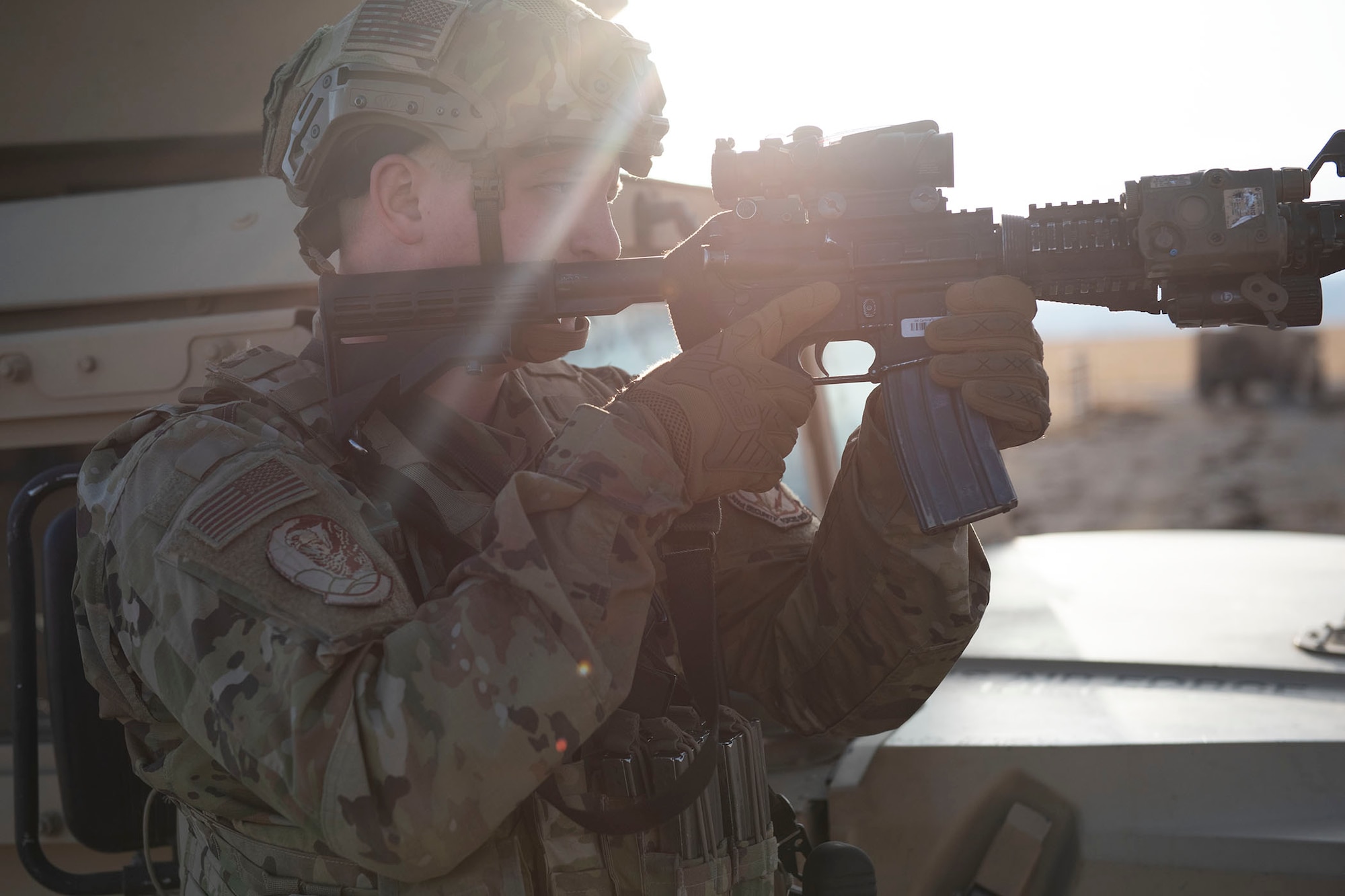 A defender stands with his rifle raised looking towards the right side of the fame. He is pointing down range, sun is shining brightly in the background of the image.