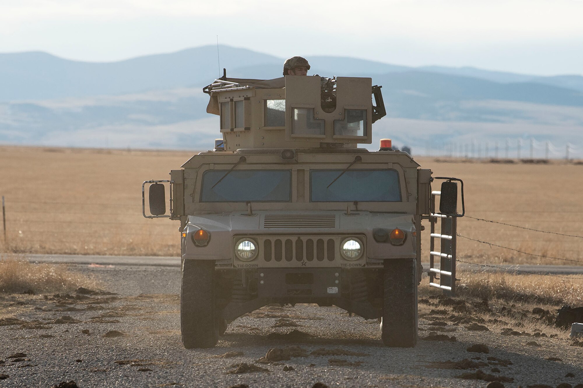 A humvee sits in the center of the road driving towards the entrance of the launch facility with a blue mountain range in the background of the photo.