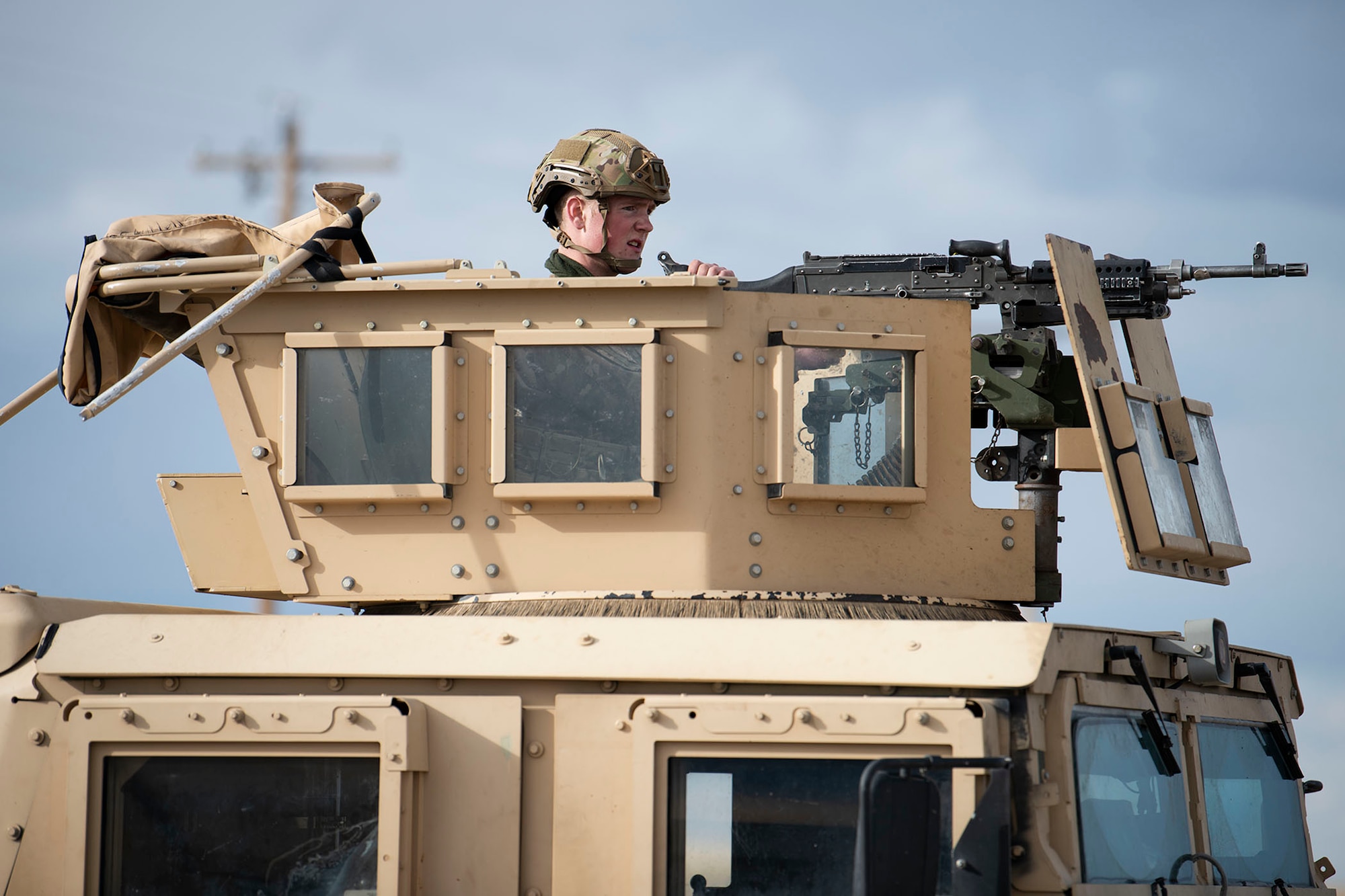 Defender sits in the turret of Humvee and looks towards the right side of the frame towards the distance.