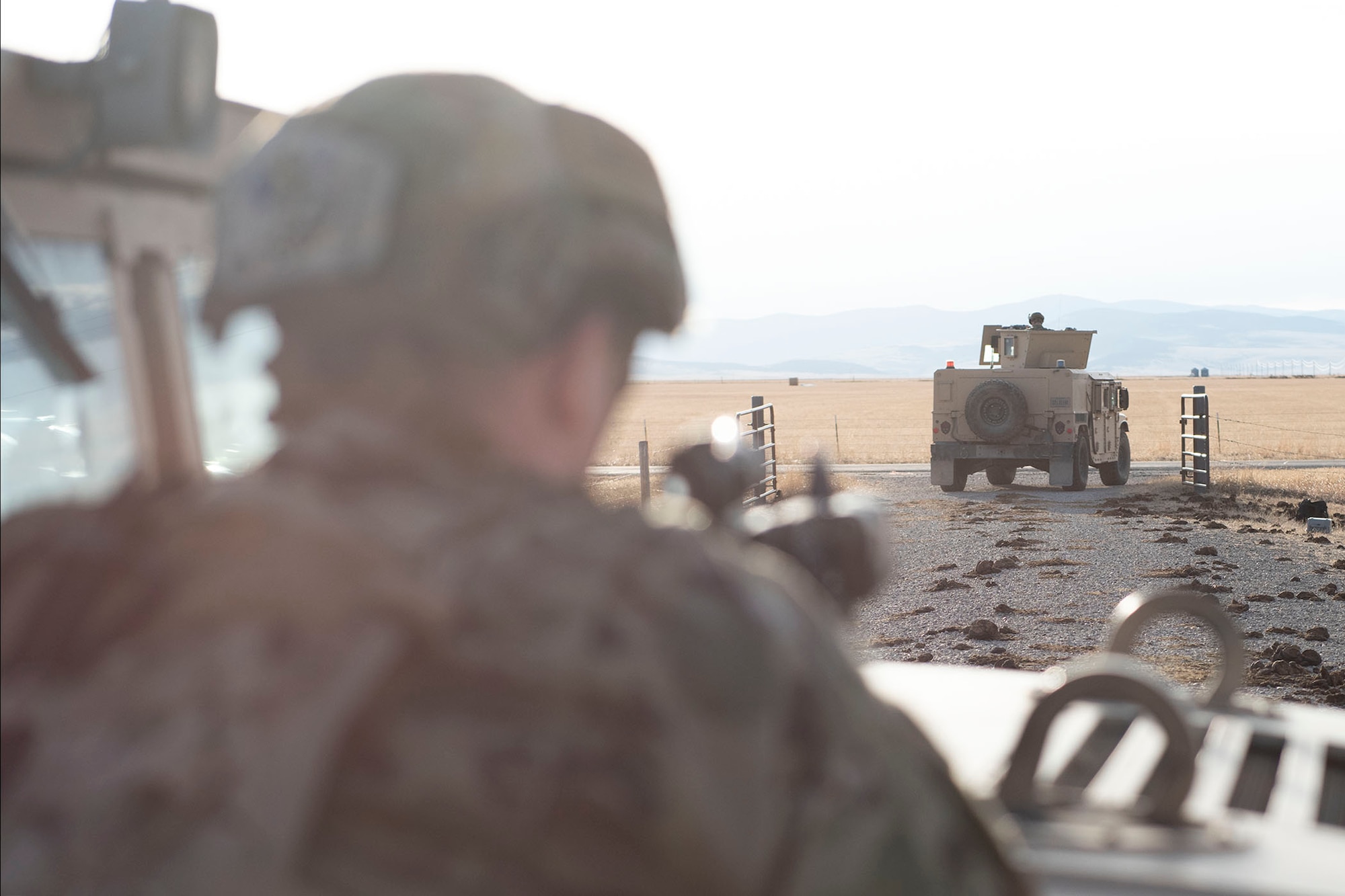 A defender leans against a humvee with his rifle pointing out in front of him (blurred) and in the forefront is a humvee driving off into the distance