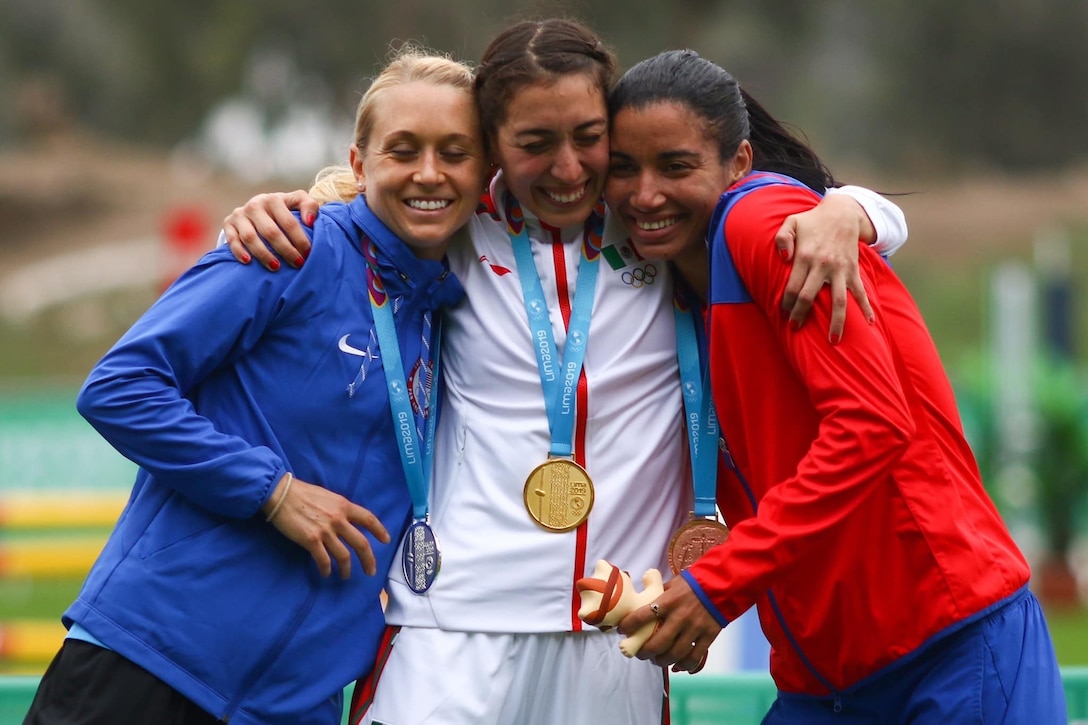 Three women wearing sports uniforms and medals around their necks hug one another.