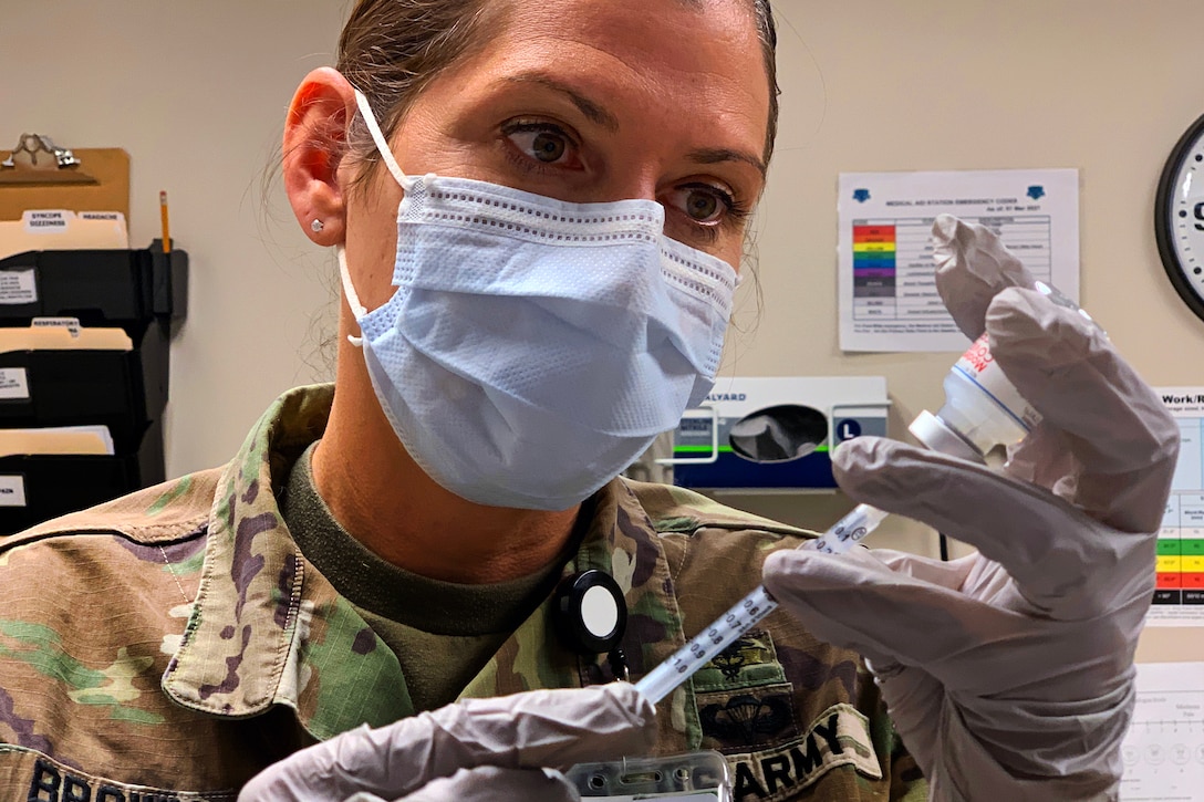 A soldier wearing a face mask and glove holds a syringe while inserting it into a vial.