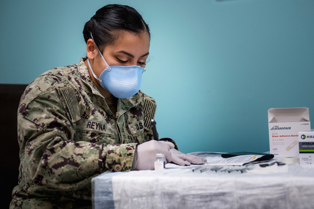 A sailor wearing a face mask and gloves sits at a table filling out paperwork.