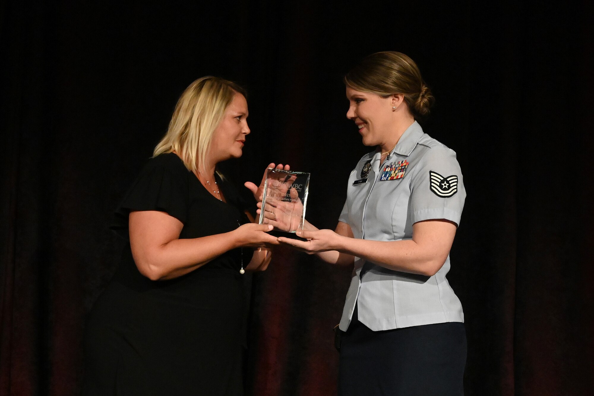 A female Airman in short-sleeve blue military uniform accepts and award from another woman in a black dress in front of an audience at the Holiday Inn, Fargo, N.D., March 30, 2021.