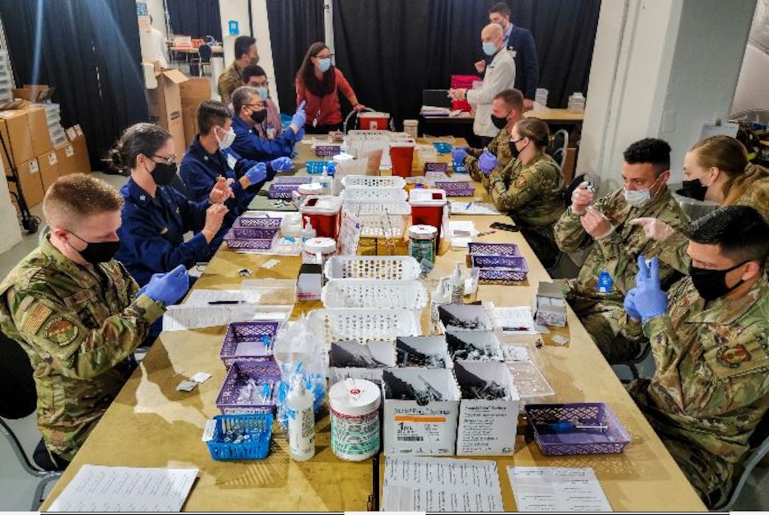 Eight male and female airmen and one civilian, who are all wearing face masks and gloves, sit at a long table cluttered with plastic bins and syringes; several of the airmen hold fill syringes with the contents of small vials. Three other people stand near the end of the table.