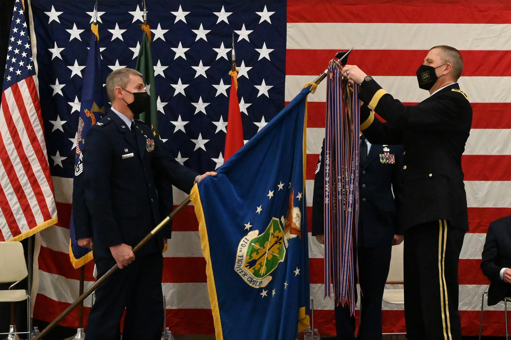 Maj. Gen. Al Dohrmann, the N.D. adjutant general places a symbolic streamer on the end of a flag staff being held by Col. Darrin Anderson, the 119th Wing commander during a ceremony on a stage in front of a large American flag at the North Dakota Air National Guard Base, Fargo, N.D., March 6, 2021.