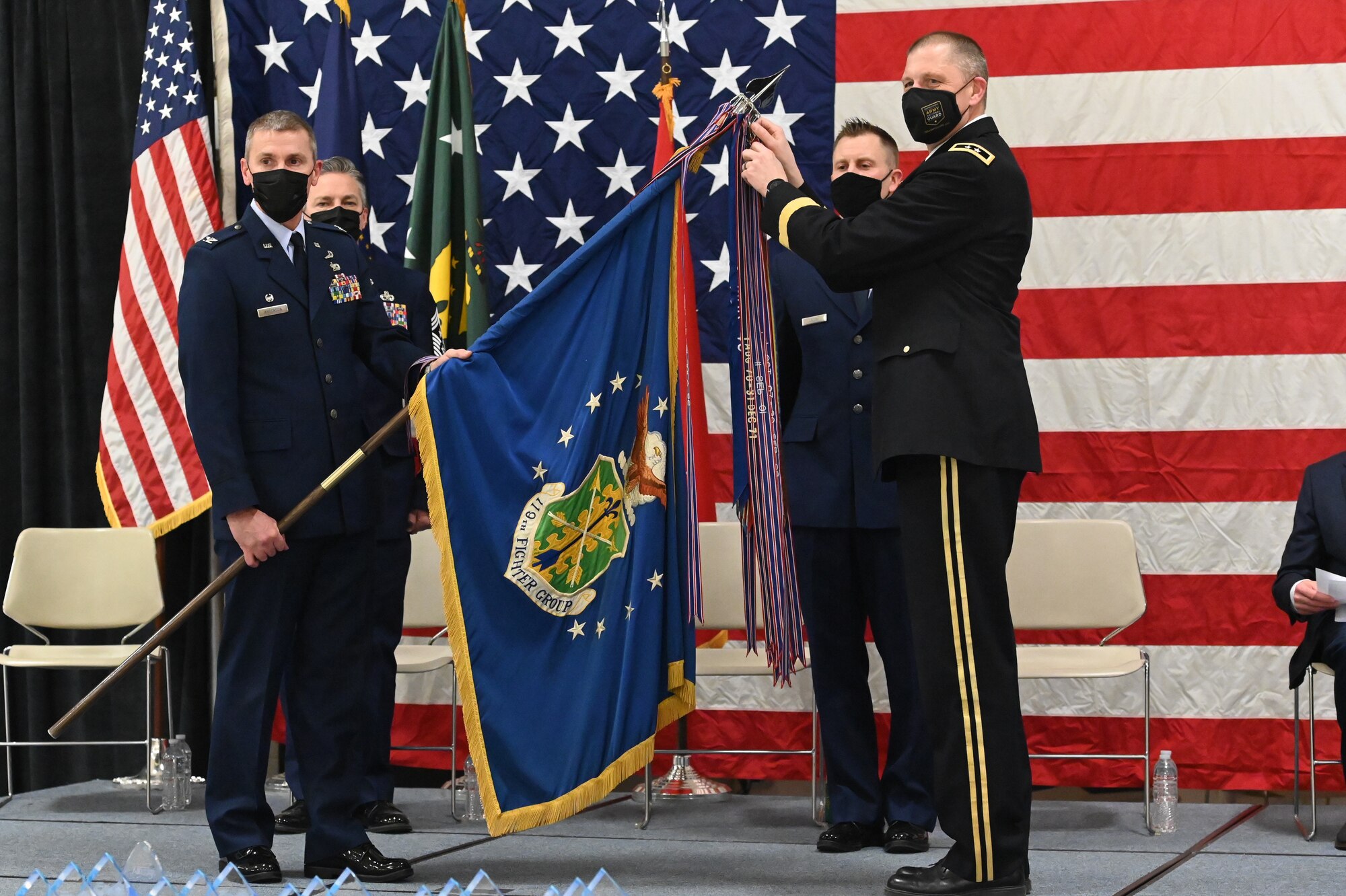 Four military members in uniform stand on a stage in front of a large American flag holding a flag staff to place a symbolic streamer representing the unit accomplishment of earning it's 22nd Air Force Outstanding Unit Award at the North Dakota Air National Guard Base, Fargo, N.D., March 6, 2021.