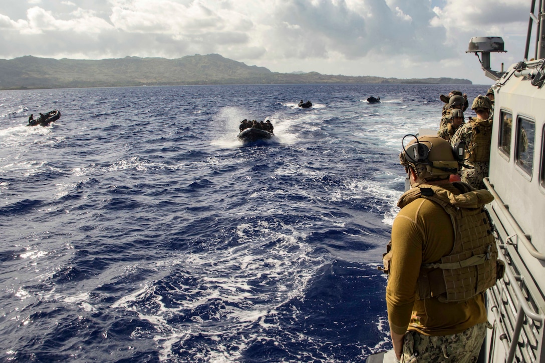 Service members standing on a ship observe service members riding in small boats.