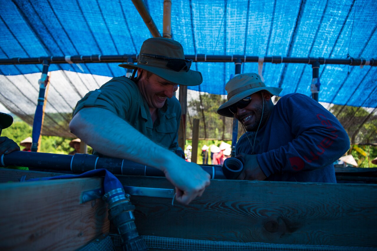 Two men work to manipulate a pipe that's attached to a wooden box while standing under a tarp in an outdoor work.