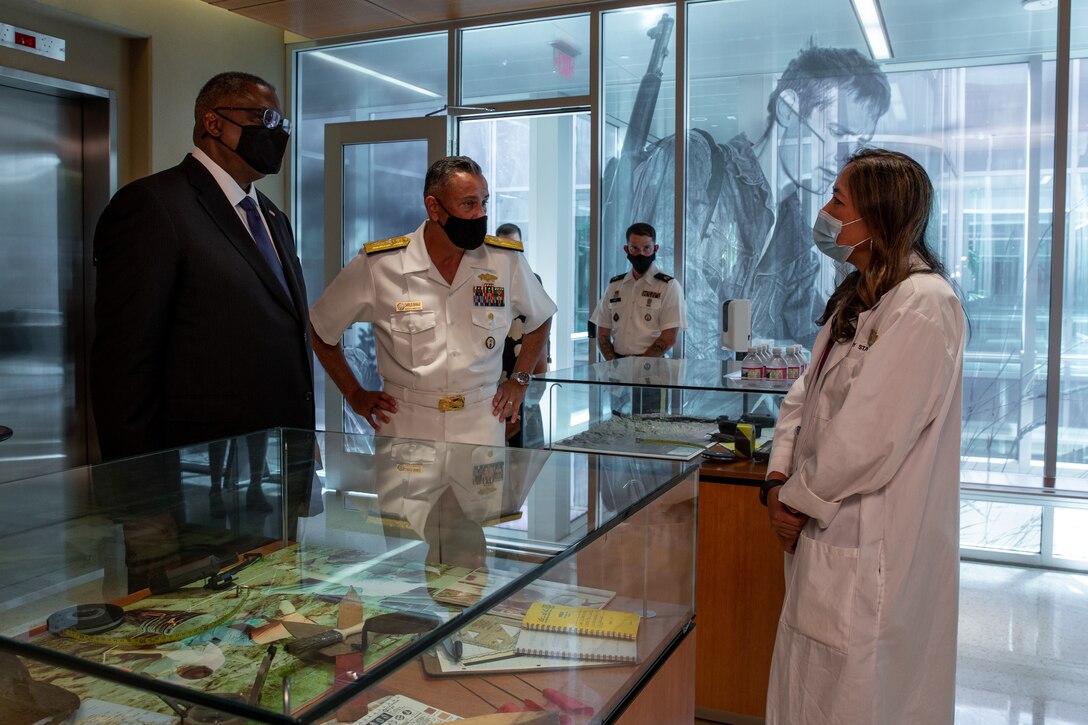 A man in a business suit and a man in a military uniform talk to a woman wearing a lab coat.