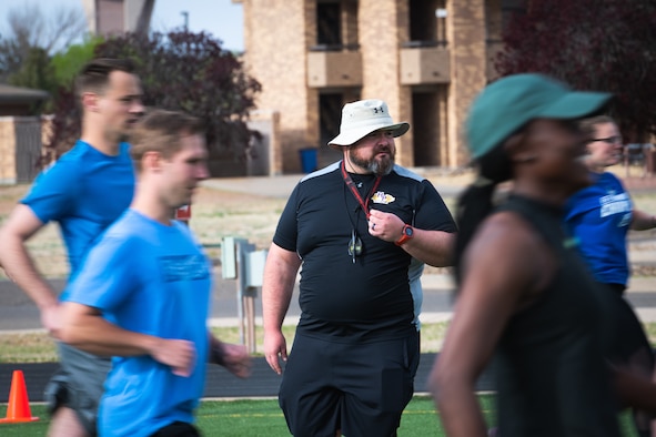 Man in a black shirt, black shorts and a wide-brim bucket hat watches others run around him on a track.