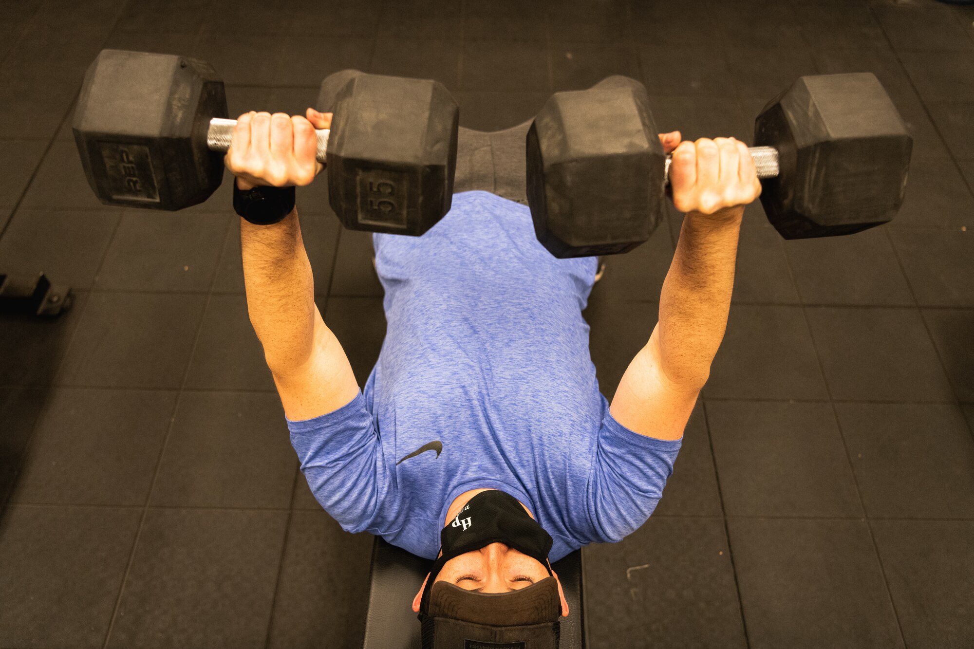 Man in a purple shirt, black hat and black mask lifts two weights off of his chest.