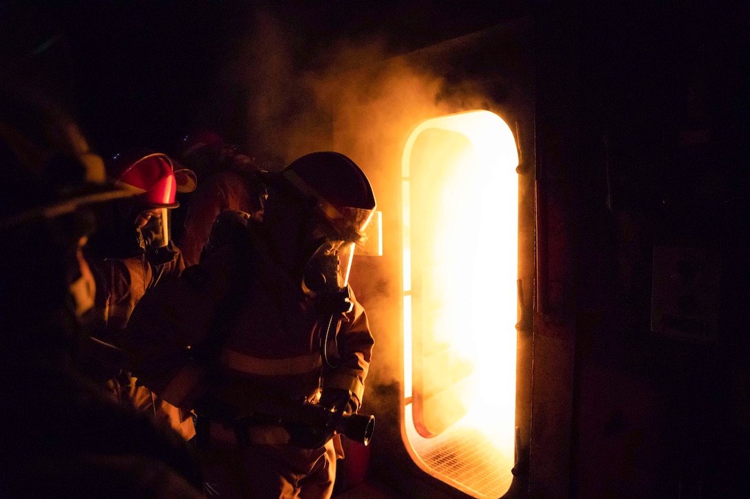 Sailors stand in a dark area in front of a door that opens up to a room engulfed in fire.