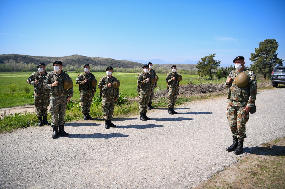 Eight men in military uniforms stand in formation on a gravel road. In the background are mountains.