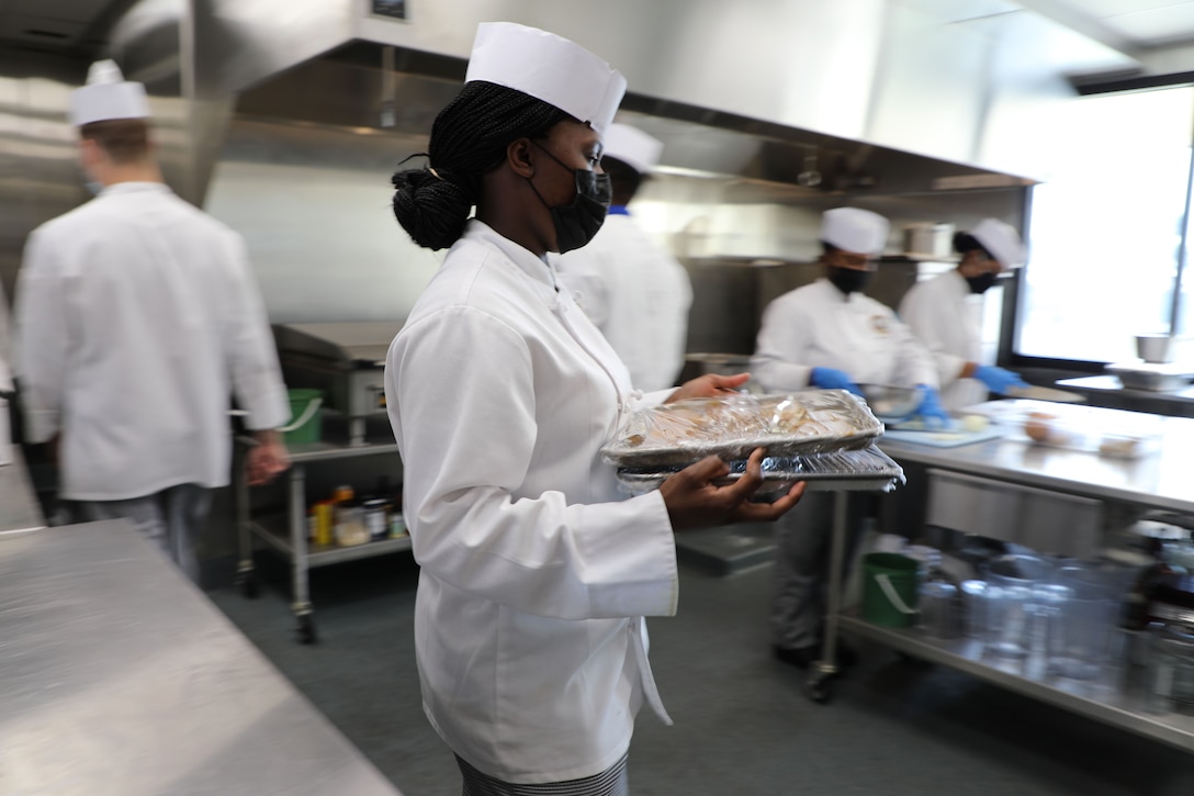 A sailor carries trays of food in a kitchen as others prep food nearby.