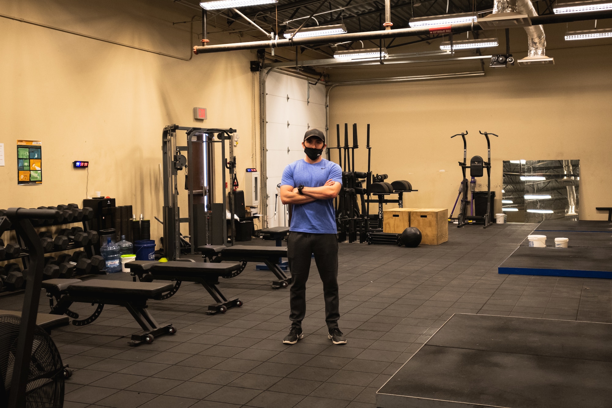 Man in a purple shirt, black pants, black hat and black mask stands in a large, dimly lit room surrounded by gym equipment.