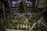 A military member grasps cargo netting over a pallet of green cylinders.