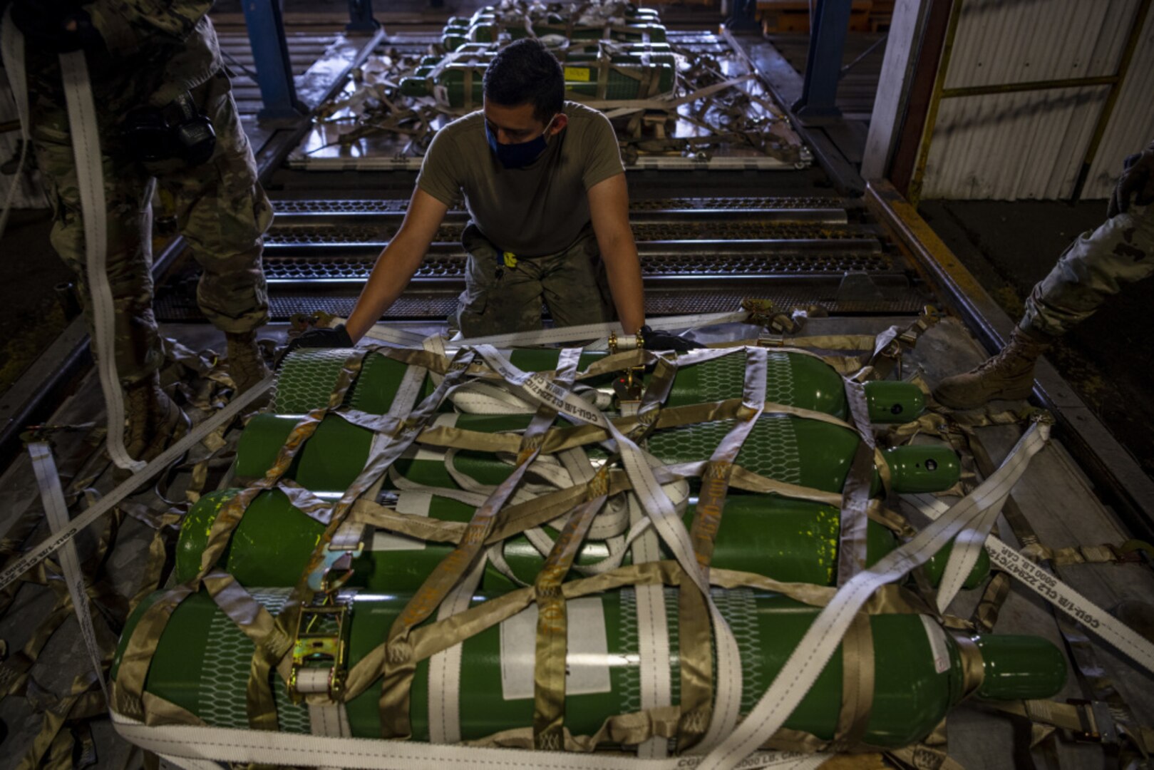 A military member grasps cargo netting over a pallet of green cylinders.