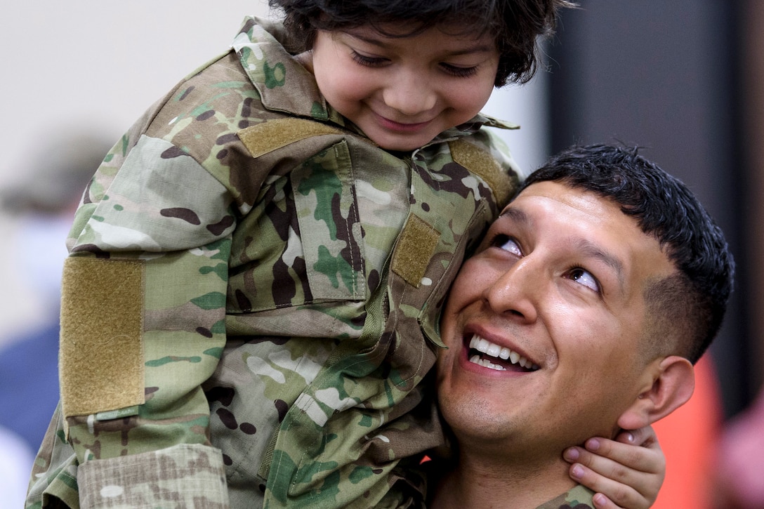 A soldier looks up and smiles at a smiling boy perched on his shoulder.