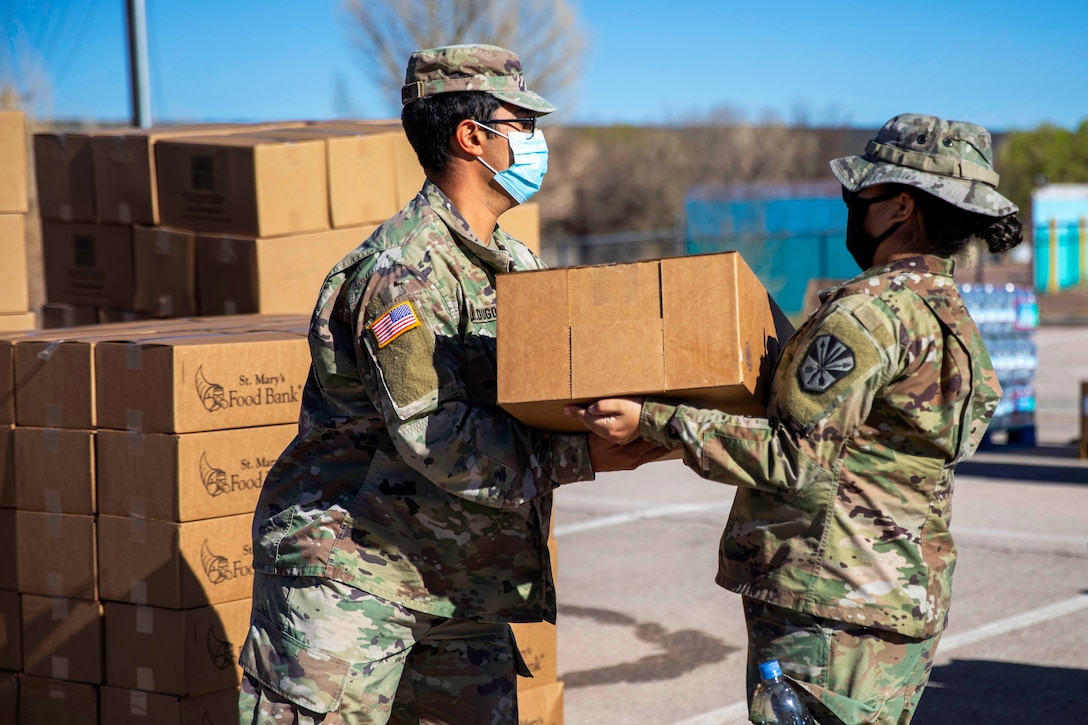 Two soldiers wearing face masks carry a box of food and supplies at a drive-thru food bank­­.