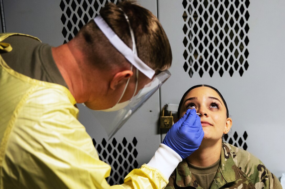 A medical technician wearing personal protective equipment swabs the nose of a soldier sitting in a chair.