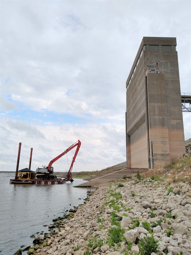 Dredging equipment is currently operating from a floating barge positioned in front of the outlet works tower.