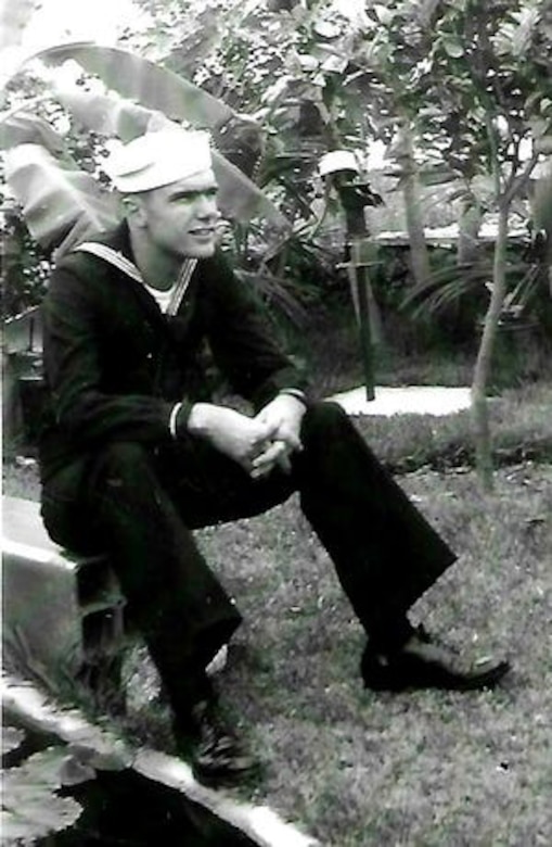 A man in a sailor's uniform sits outside in a tropical setting with his forearms resting just above his knees.
