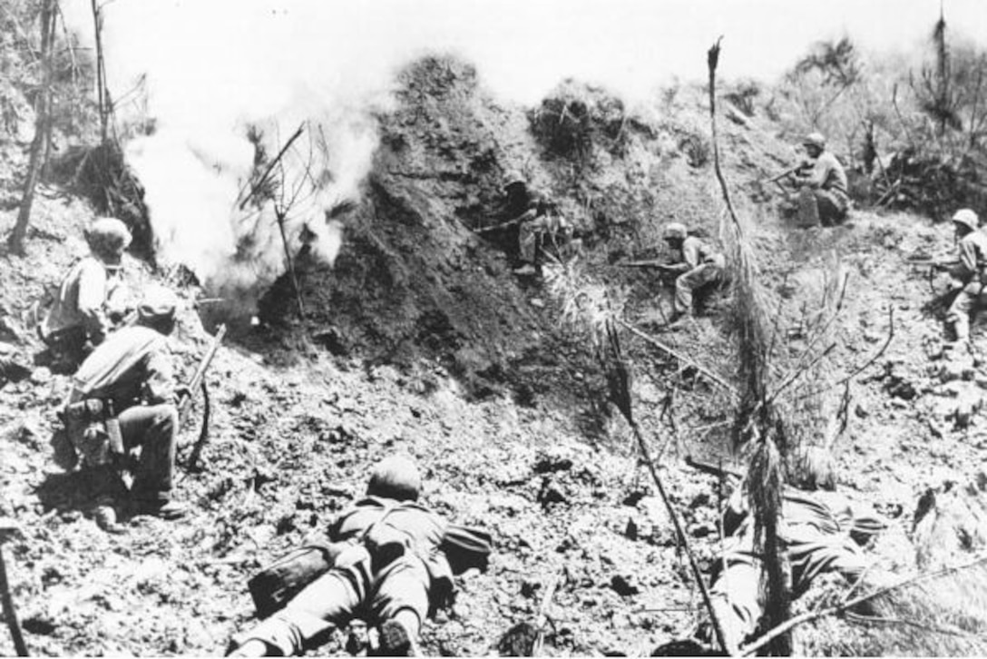 Men with rifles crouch several feet from a hillside cave entrance that has smoke coming out of it.