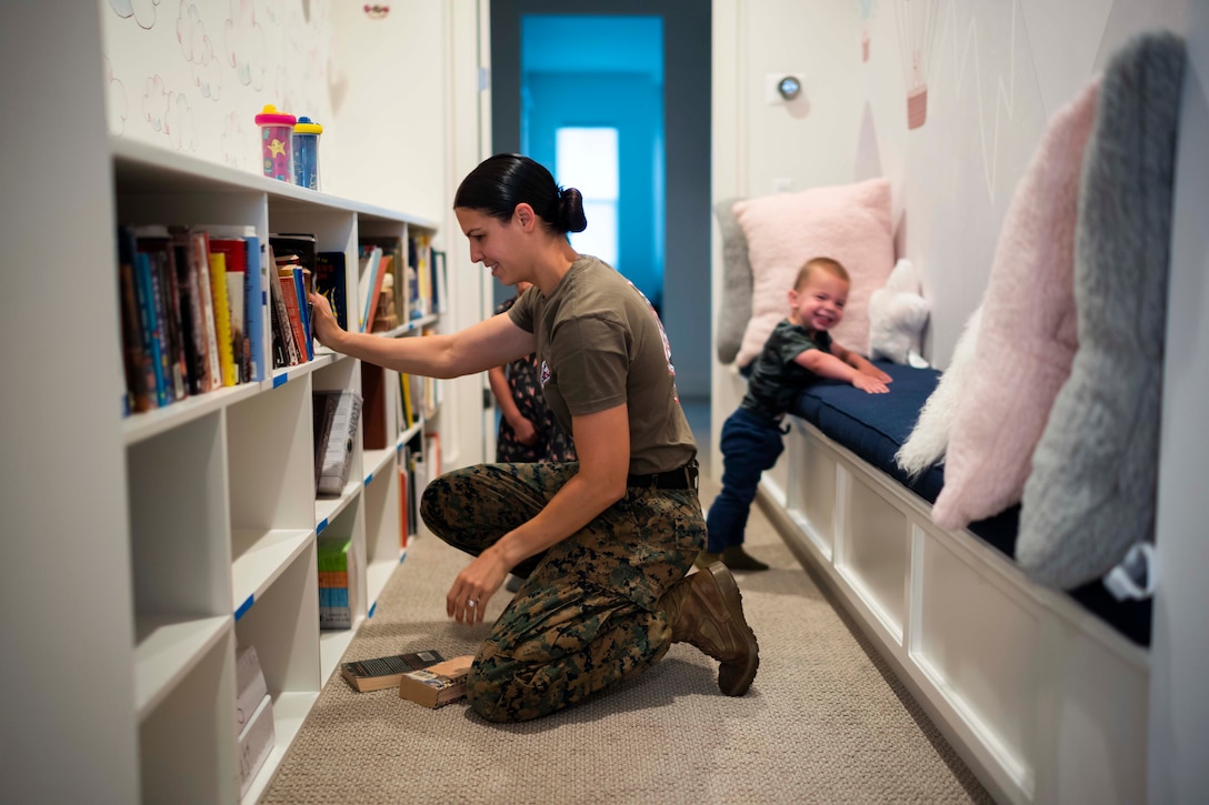 A Marine kneels next to a bookcase as two children stand behind.