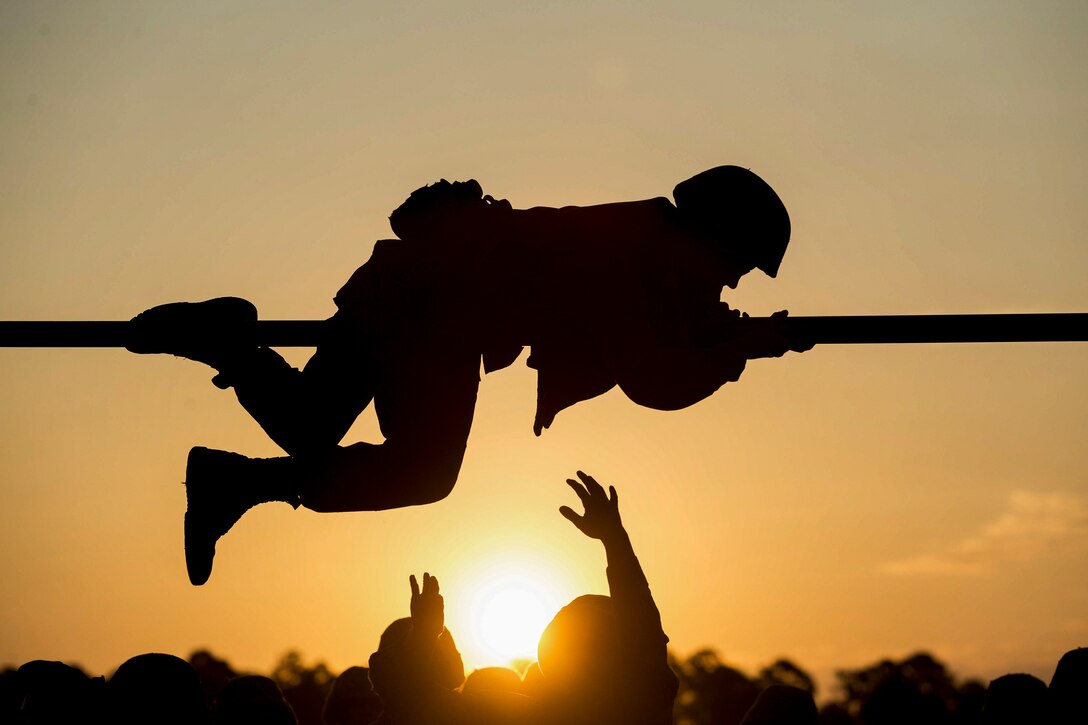 A Marine Corps recruit, shown in silhouette, crawls on a horizontal pole above the ground as fellow recruits hold their arms up.