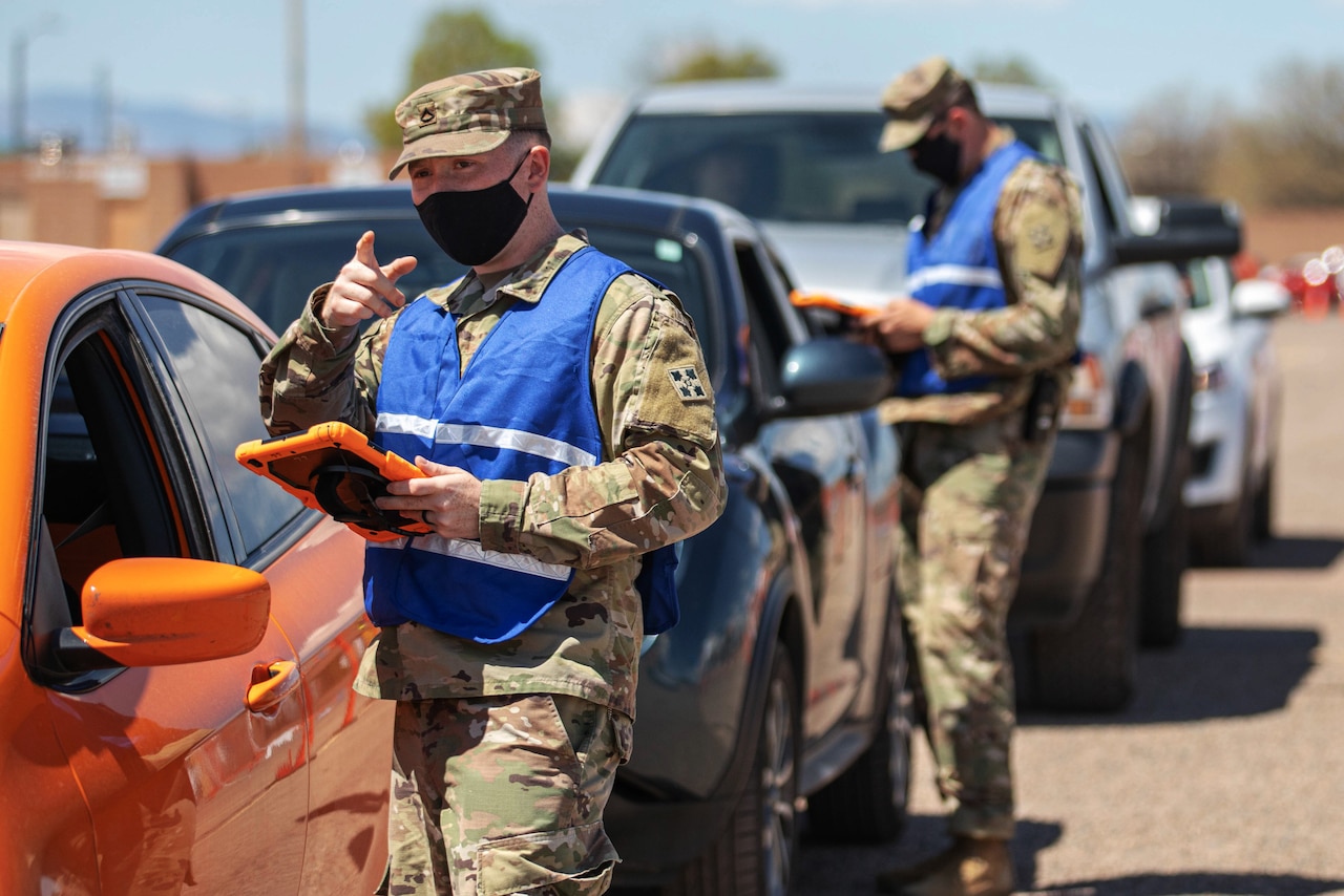 A soldier wearing a face mask gives directions to a community member in their car.