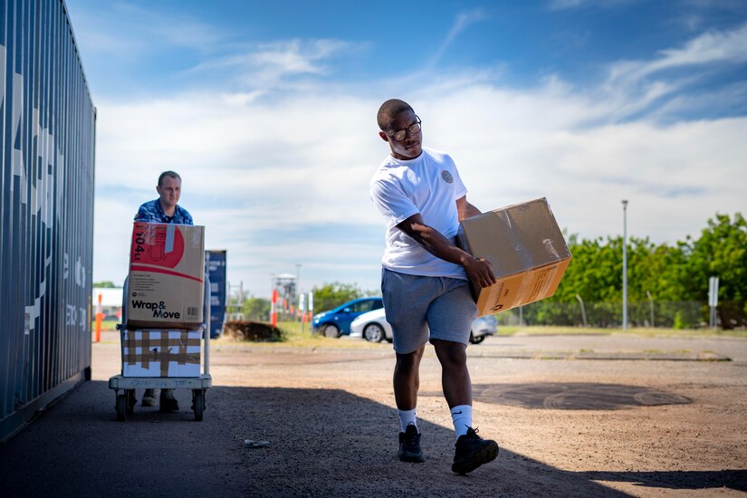 One man pushes a cart loaded with boxes while another man carries a box.