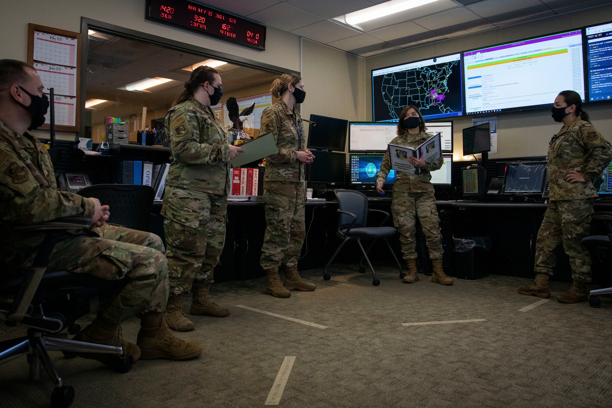 Airmen stand next to large screens, one Airman holds a binder.