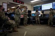 Airmen stand next to large screens, one Airman holds a binder.
