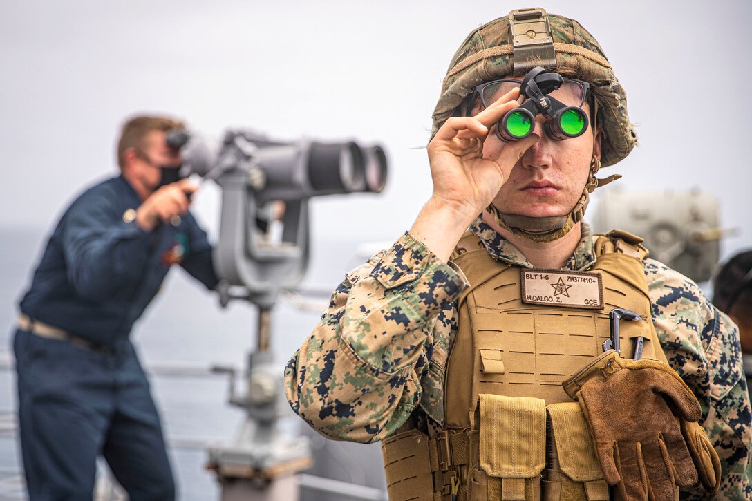 A Marine looks through small binoculars in the foreground while a sailor looks through big binoculars in the background on a ship's deck.