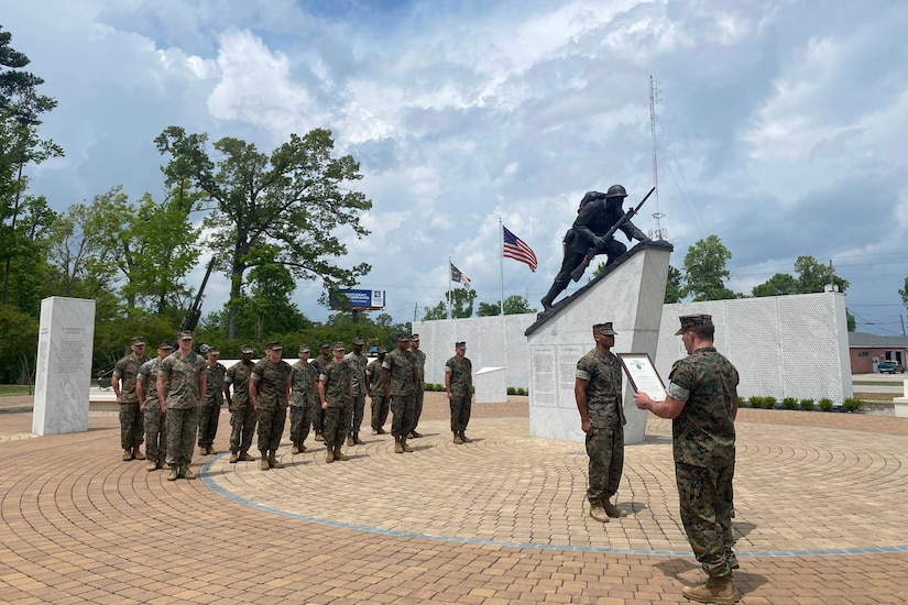 Standing next to a statue outdoors, a Marine holds a plaque as he faces another Marine; other Marines watch.